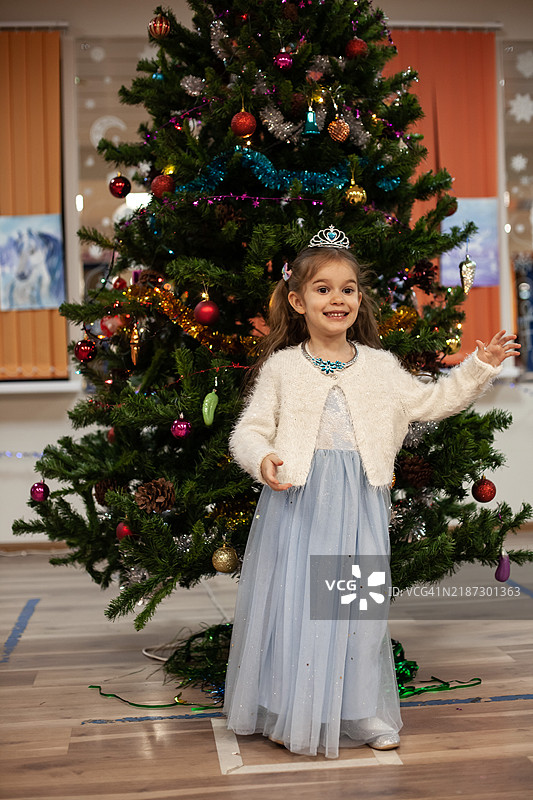 一个3-4岁的小女孩穿着美丽的蓬松蓝色裙子，戴着皇冠，背景是一棵圣诞树。图片素材