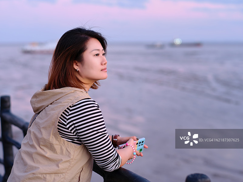 在日落时分，一位东亚美女倚靠在木栅栏上，凝视着远方。图片素材