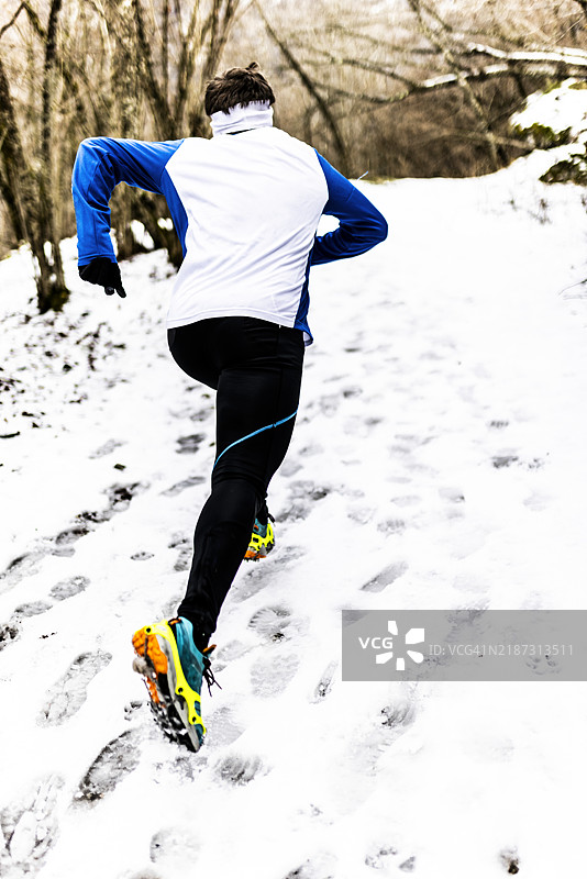 年轻男子在雪山的森林小径上跑步训练，穿着冬季服装，手套和鞋子上带有微型冰爪。图片素材