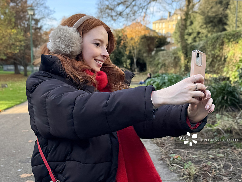 特写镜头，迷人的红发女性穿着黑色羽绒服、阿兰毛衣、红色肩包和围巾，戴着仿毛皮耳罩，手持智能手机自拍，背景为公共公园，重点在前景。图片素材