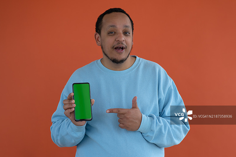 一位快乐、笑着的非裔美国男子手持手机，指着手机，站在简单的背景前。图片素材