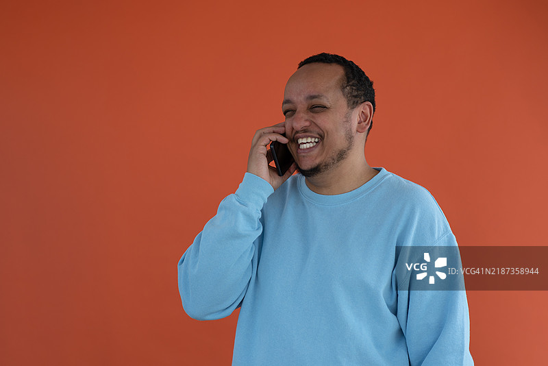 一位快乐的非裔美国男子微笑着，手持手机靠在耳边，正在通话，站在简单的背景前。图片素材