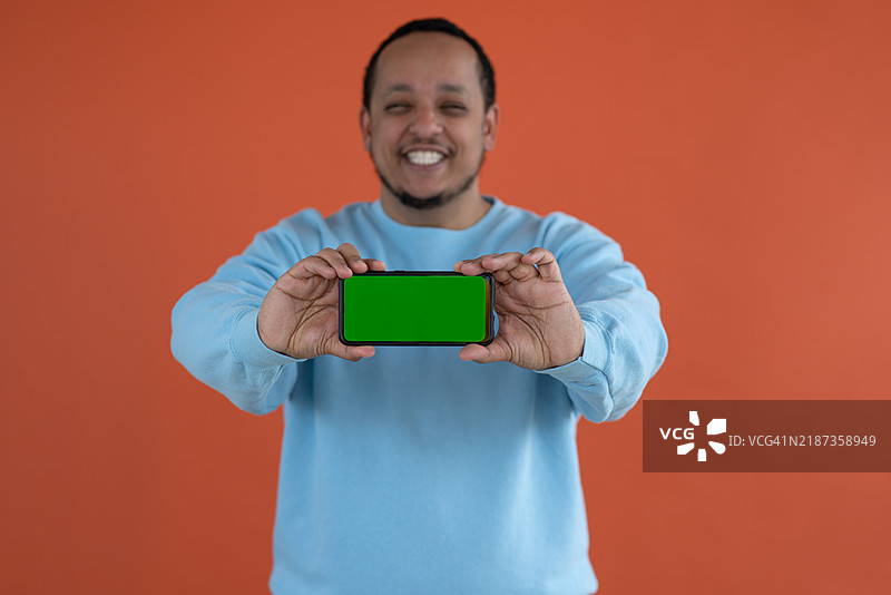 一位年轻、快乐的非裔美国男性伸出手，手中握着一部手机，站在简单的背景前。图片素材