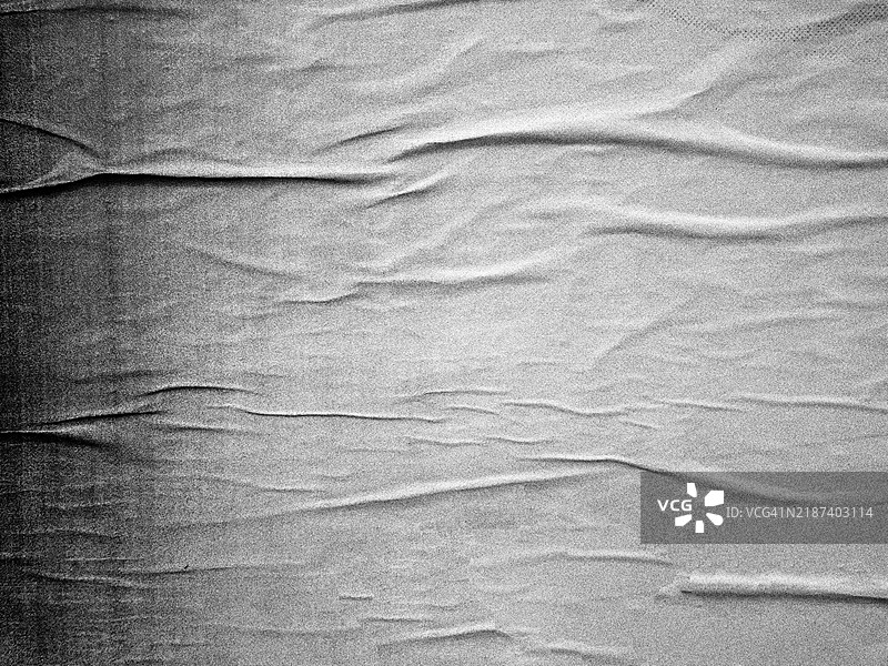 墙上贴着一张皱褶的灰色海报的特写。粗糙的纹理。黑白照片带有噪点效果。阳光照射。图片素材