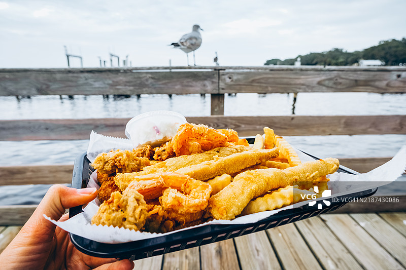 美味的海鲜拼盘，包含炸虾和鱼，位于海边的木栈道上。快餐海鲜。餐厅内的一次性餐具中盛放着炸虾、鱼、薯条和柠檬片。图片素材