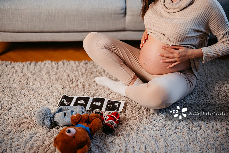 年轻孕妇坐在客厅地板上，旁边是婴儿衣物、超声波照片和一只毛绒泰迪熊。图片素材