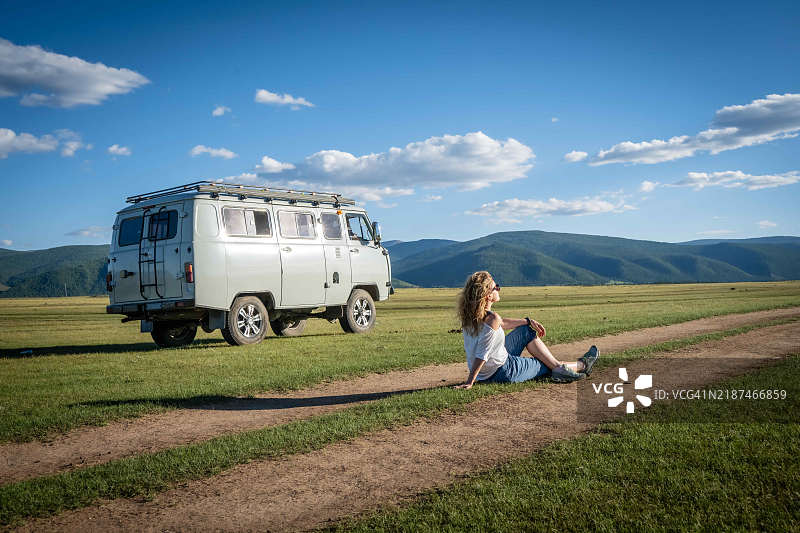 一位女性坐在乡村道路上放松，目光投向远方，享受在蒙古的周末公路旅行。图片素材