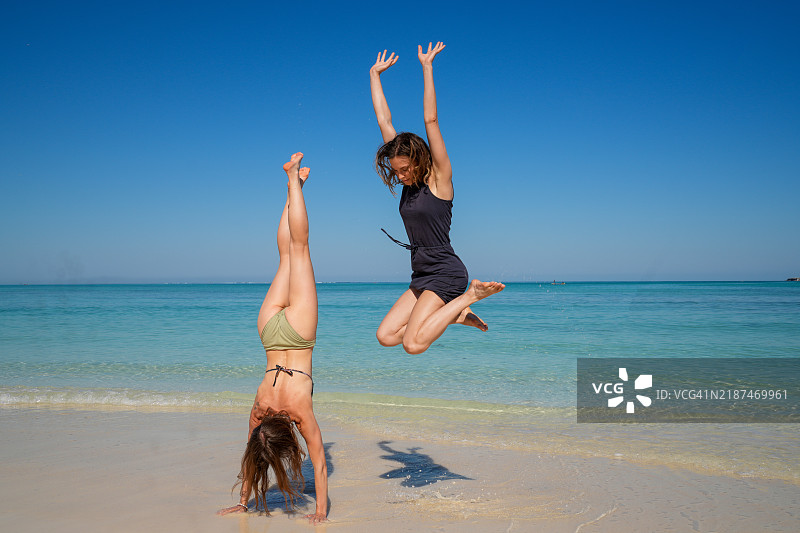 两位年轻女性在马达加斯加的白色沙滩上跳跃，一位举起双手，另一位倒立。图片素材