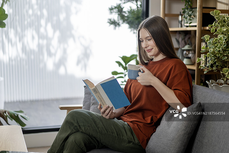 女人坐在舒适的沙发上阅读一本书，喝着咖啡或茶。图片素材