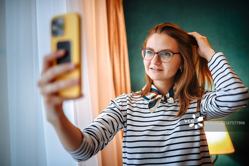 在她舒适的卧室里，一位穿着条纹毛衣的女性正在试戴丝巾并与造型师咨询，同时拍摄自拍照。图片素材