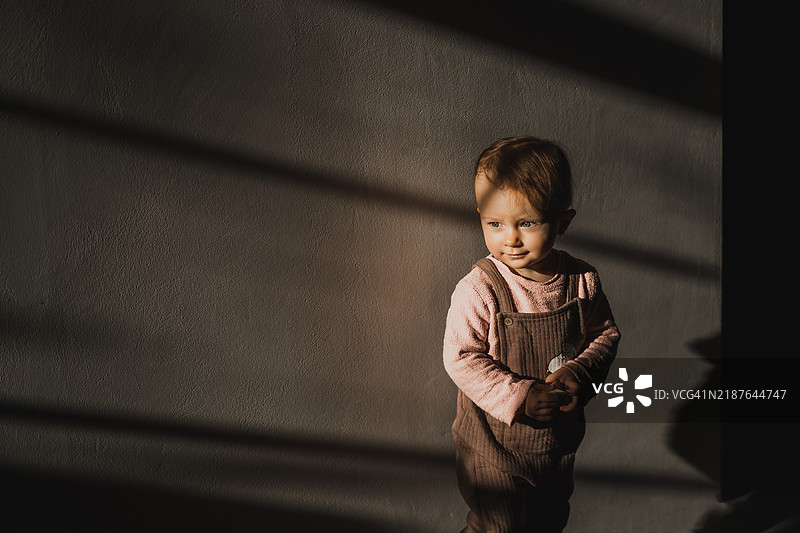可爱的1岁女孩站在混凝土墙旁，光线柔美，目光投向远方。图片素材