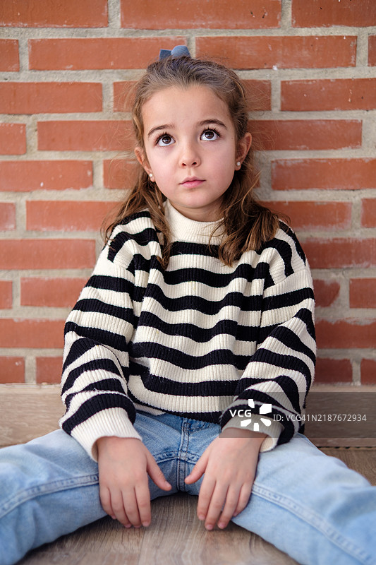 沉思的孩子坐在地板上仰望，背景是砖墙图片素材