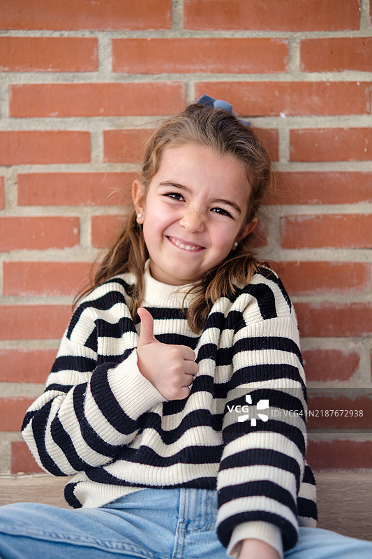 微笑的女孩在砖墙背景前做出竖起大拇指的手势图片素材