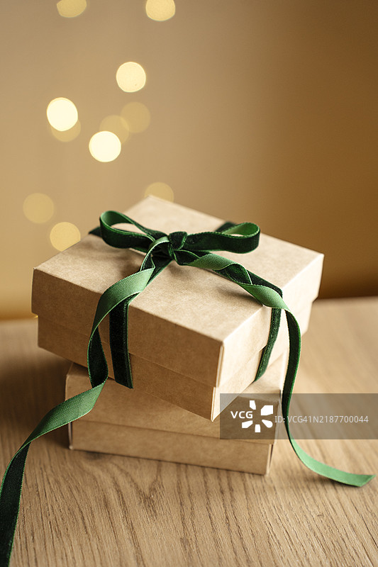 两只棕色礼品盒叠放在木桌上，系着绿色天鹅绒蝴蝶结和丝带，背景是模糊的灯光散景。图片素材