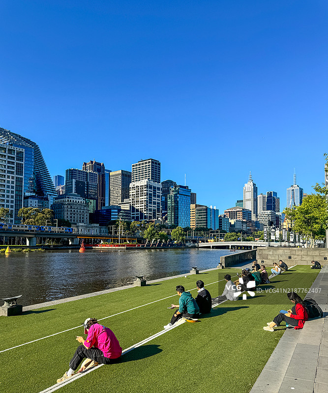 年轻人在墨尔本市中心天际线附近的雅拉河旁的草地上坐着图片素材