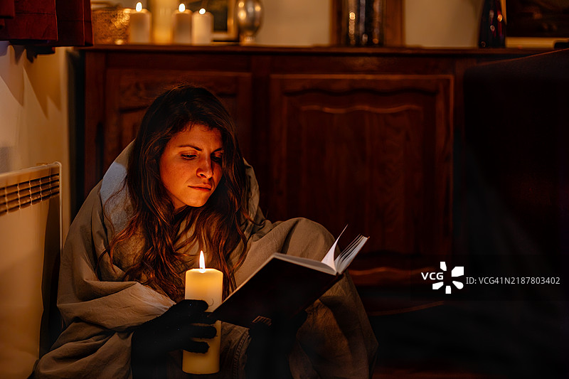 一位女性坐在地板上，裹着毯子，在停电期间的温馨家中，借着烛光阅读一本书。图片素材