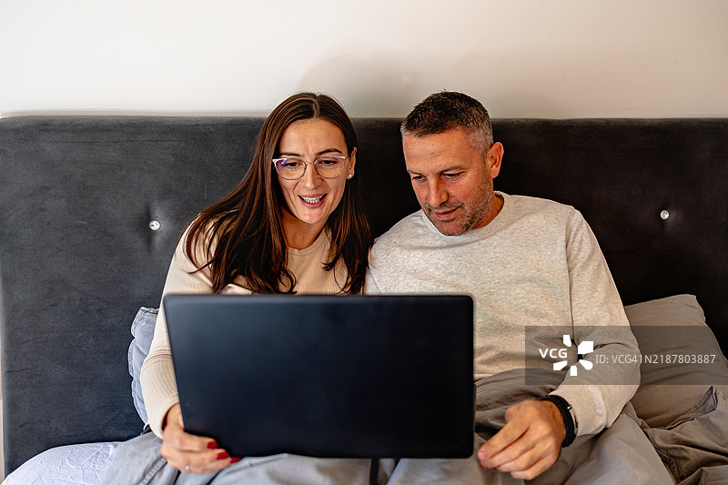 一位年轻女性和一位男性在舒适的室内空间中，悠闲的早晨一起坐在床上共享一台笔记本电脑。图片素材