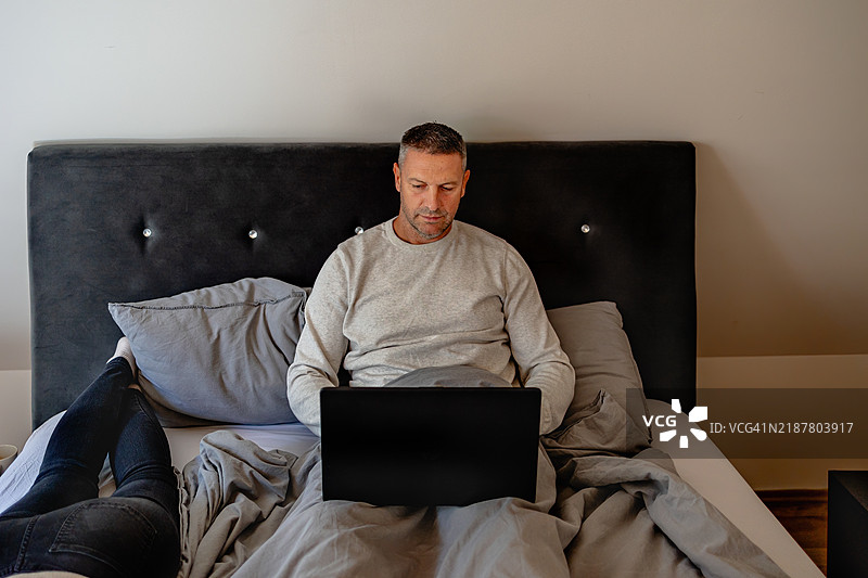 年轻男子在温馨的卧室里坐在床上使用笔记本电脑，正值下午。图片素材