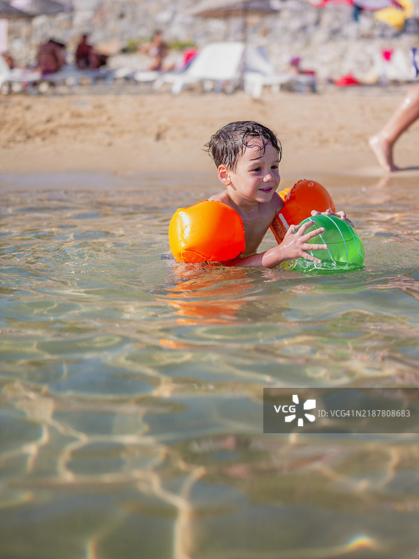 孩子们在海边玩球。一位漂亮的男孩在海岸上玩耍。他穿着充气游泳袖。6岁。夏天的白天。图片素材