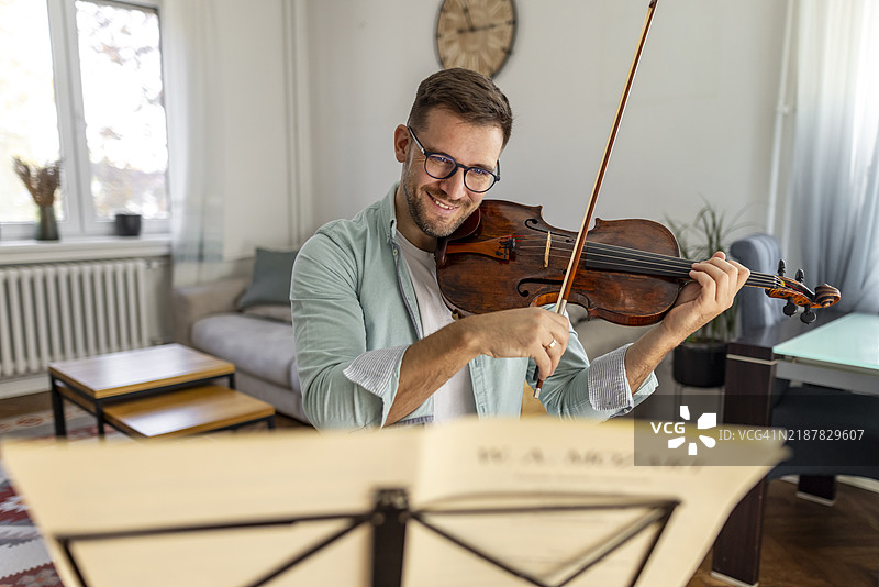 微笑的男人在舒适的客厅里拉小提琴，旁边有乐谱图片素材