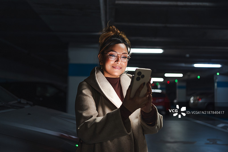 微笑的拉丁裔女商人在昏暗的停车场查看手机，捕捉到城市中一刻的联系与私密。图片素材