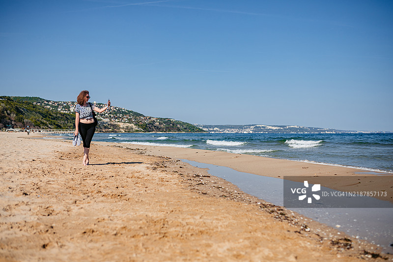 年轻女性在保加利亚阿尔贝纳的海滩上走路时用手机自拍图片素材