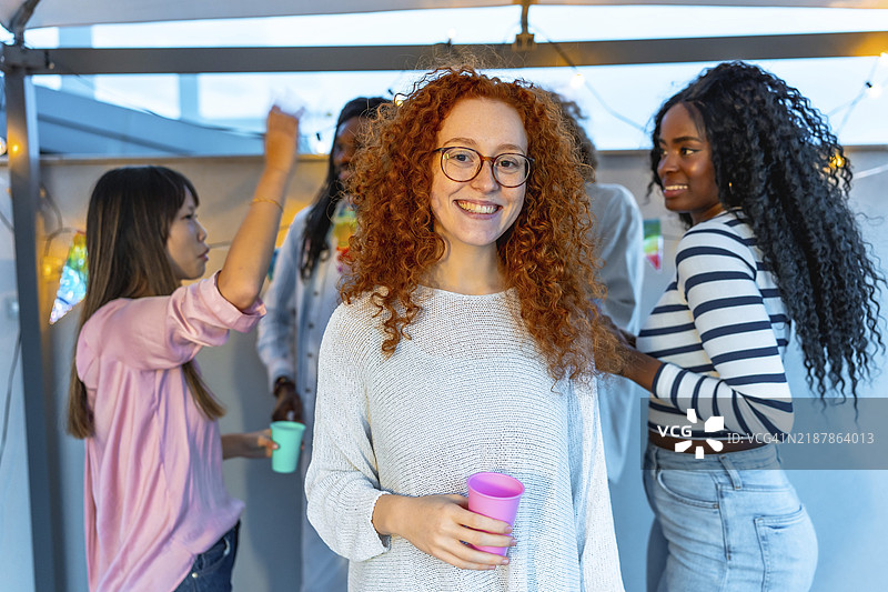 年轻女性，红色卷发，戴着眼镜，微笑着站在屋顶派对上，手中拿着啤酒杯，身边是多种族的朋友们。图片素材
