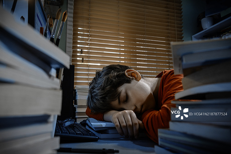 疲惫的学生趴在书桌上睡觉，周围是书籍和电脑。图片素材