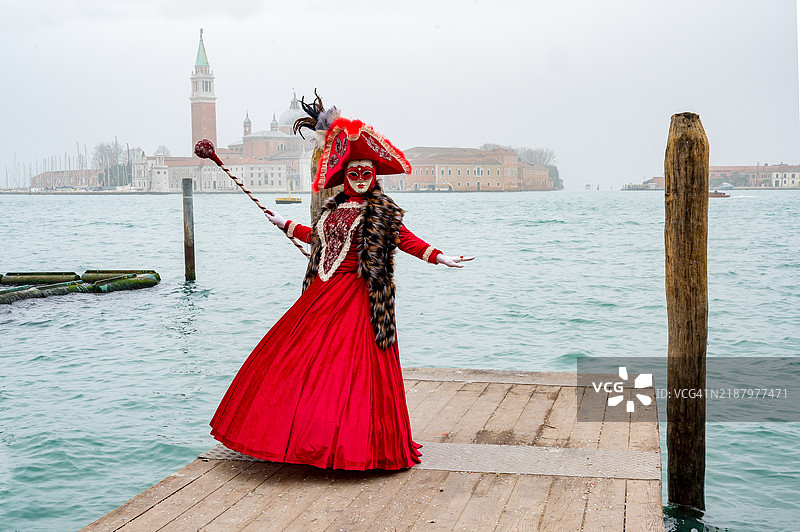 身穿红色裙子的面具女郎在威尼斯狂欢节，身后是圣乔治岛。图片素材