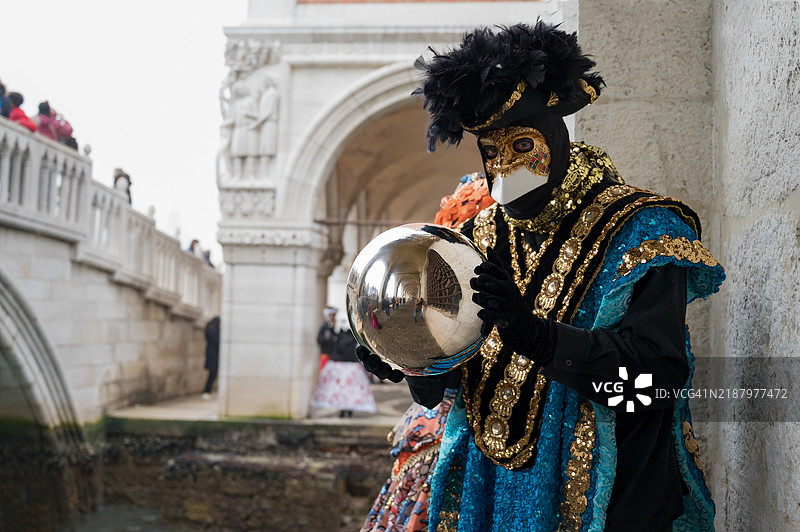 在威尼斯狂欢节上，一个戴面具的男子手持反射球，身后是总督宫。图片素材