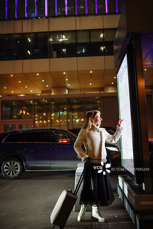 探索城市：一位女性带着行李箱在繁忙的夜间街道上与信息显示屏互动。图片素材