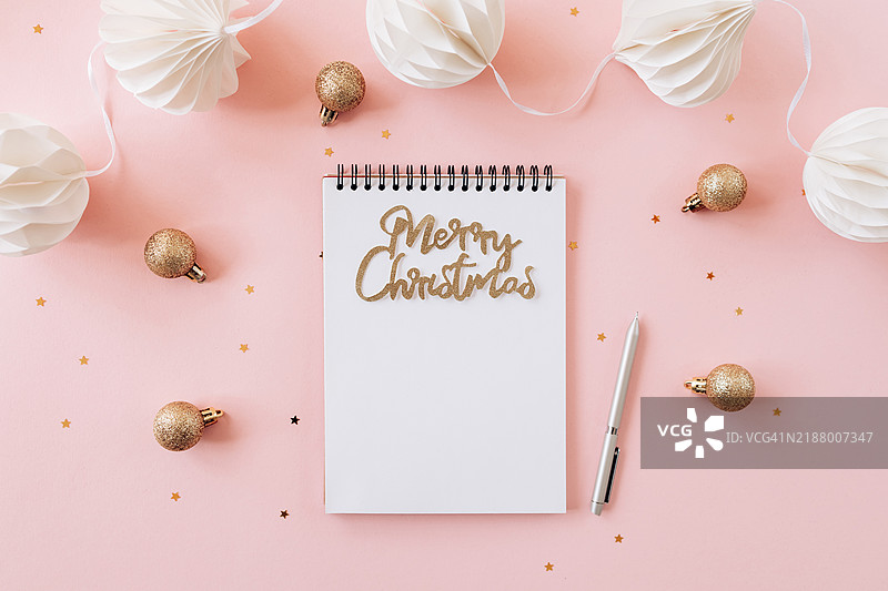 平铺构图，展示一个螺旋笔记本，上面用金色闪光字母写着“圣诞快乐”，背景为柔和的粉色。非常适合节日主题内容或节日规划。图片素材