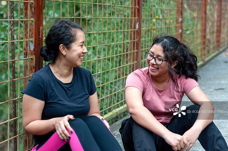 两个快乐的女性穿着运动服，坐在生锈的铁丝围栏旁，享受着锻炼时的交流与欢笑。图片素材
