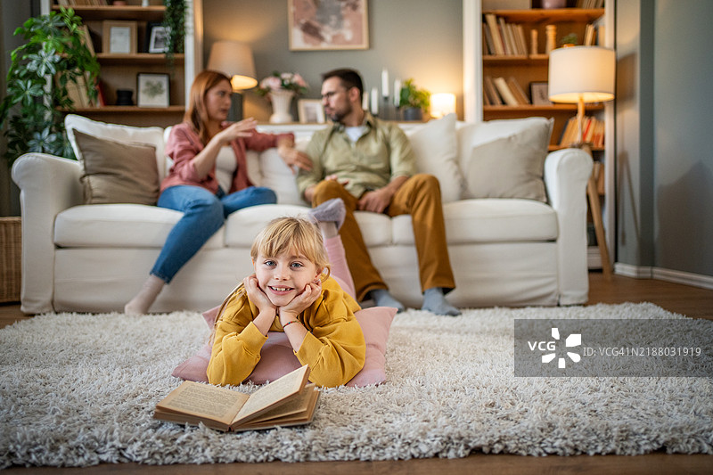 孩子躺在地板上看书，父母在沙发上交谈。图片素材