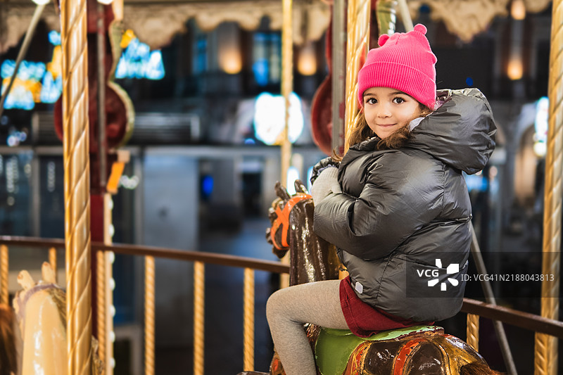 微笑的女孩戴着粉色毛线帽在游乐园的旋转木马上骑行图片素材