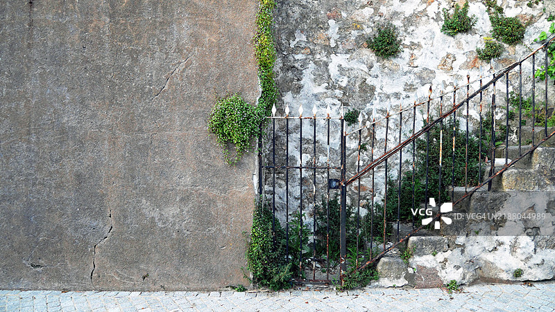 有纹理的灰色混凝土墙与古老的石阶。生锈的铁门和大门。杂草和植物。鹅卵石街道。阳光。自然色彩和风化效果。葡萄牙，波尔图。图片素材