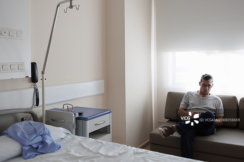 男子坐在医院病房里，旁边是一张空床。图片素材