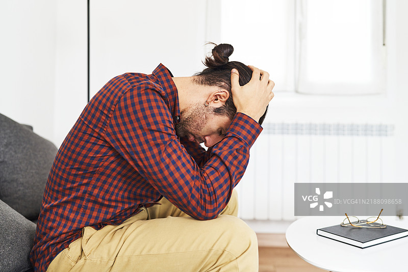 一位年轻男子穿着休闲服，面露沮丧和担忧的表情，侧坐在沙发上。图片素材