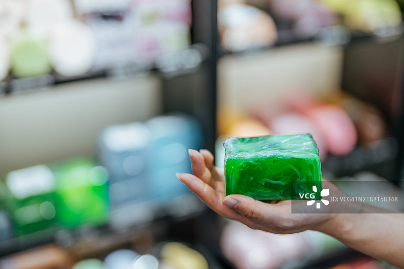 亚洲年轻女性在购买手工香皂的特写镜头。手中拿着薰衣草草本香皂，背景是各种身体护理和浴室化妆品的货架。图片素材