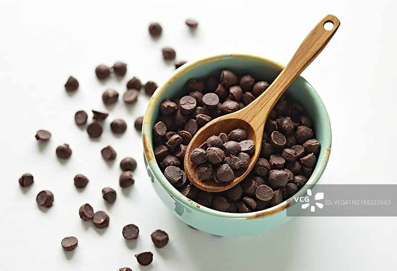 一碗巧克力豆旁边放着一把木勺，背景为白色。图片素材