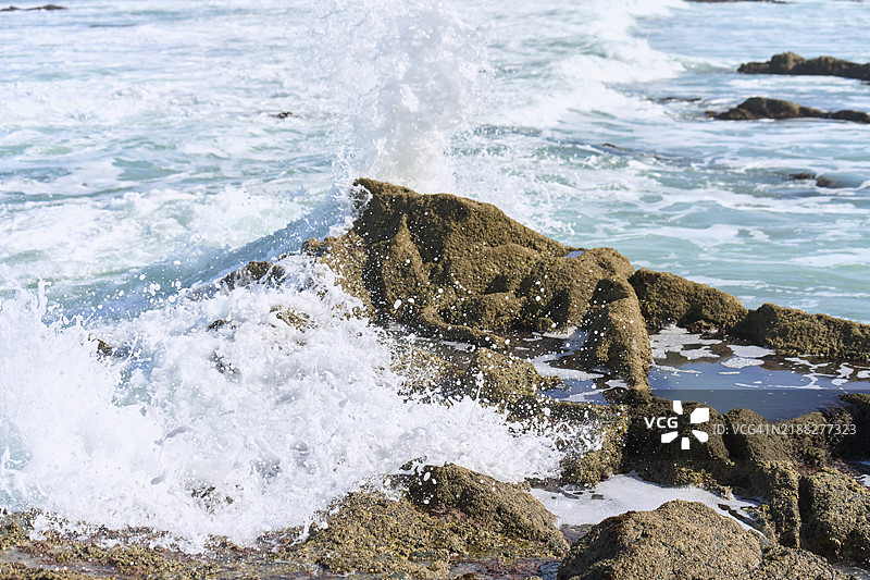 阳光照耀下，海浪在覆盖着苔藓的海滩岩石上拍打，水面上可以看到更多的岩石。图片素材
