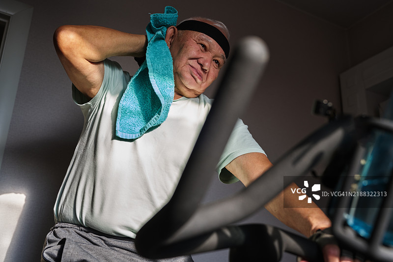 戴着头带和手套的老年男子在家中骑固定自行车进行有氧运动图片素材