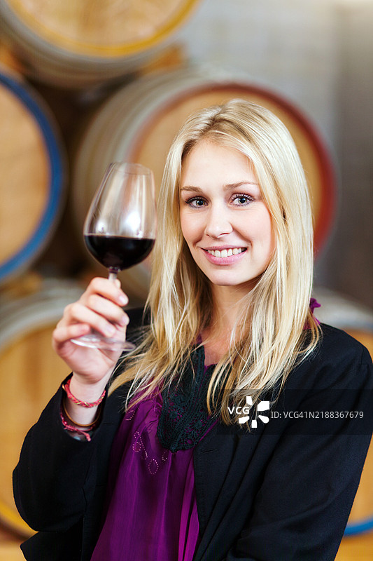 美丽的金发女人举起酒杯向你致意，背景是酒窖里的酒桶。图片素材