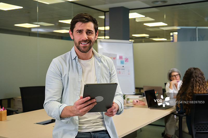 年轻的商人手持平板电脑，面带微笑地看向镜头，身处现代办公室。图片素材