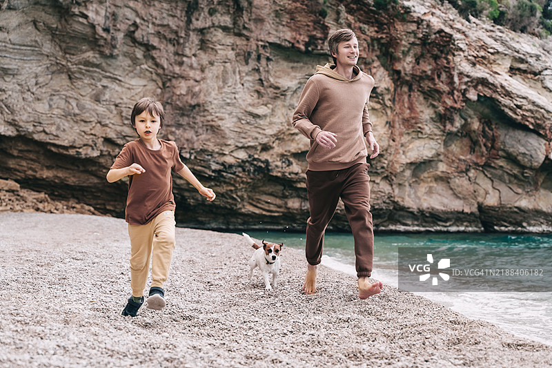 年轻父亲、小男孩和狗在沙滩上奔跑，背景是美丽的岩石纹理。图片素材