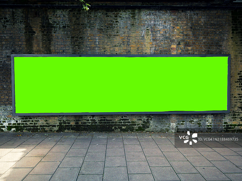 一块长而空旷的绿色矩形广告牌,位于一面风化的砖墙前。铺砌的人行道。阳光明媚。自然色彩。图片素材