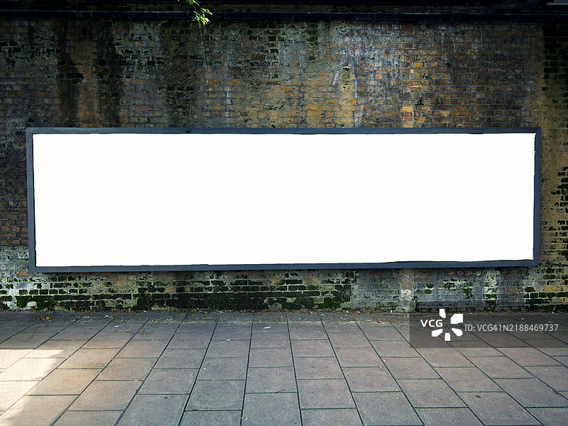 一块长而空旷的白色矩形广告牌位于一面风化的砖墙前。铺砌的人行道。阳光。自然色彩。图片素材