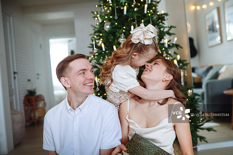 魔幻的节日氛围：父母和孩子们穿着节日服装，在明亮现代的客厅里，欢庆圣诞节，身旁是一棵美丽的圣诞树。图片素材