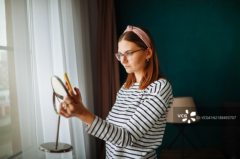 在家舒适购物：一位女性在手持镜子的情况下试戴眼镜和头带，放松地坐在她绿色墙壁的舒适卧室里。图片素材