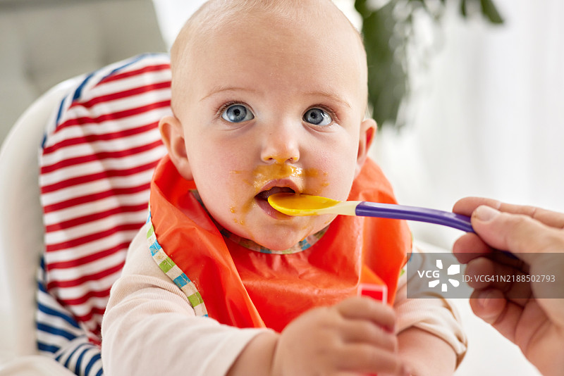 可爱宝宝在高脚椅上吃着果泥，穿着鲜艳的橙色围兜图片素材